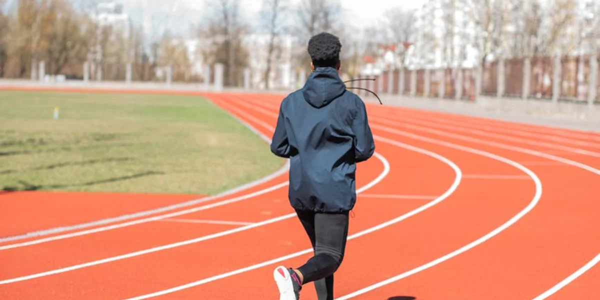 Back view of a runner on an outdoor red track, wearing a dark hoodie and pants, with a green infield and trees in the background.
