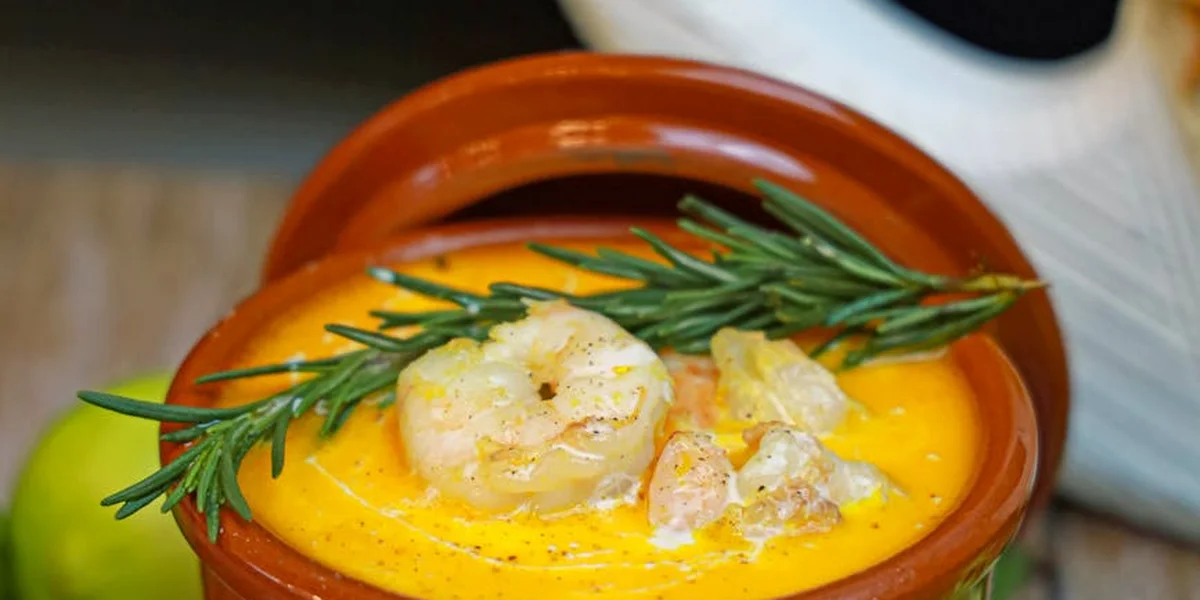 Clay pot of bright orange seafood soup topped with shrimp and a rosemary sprig