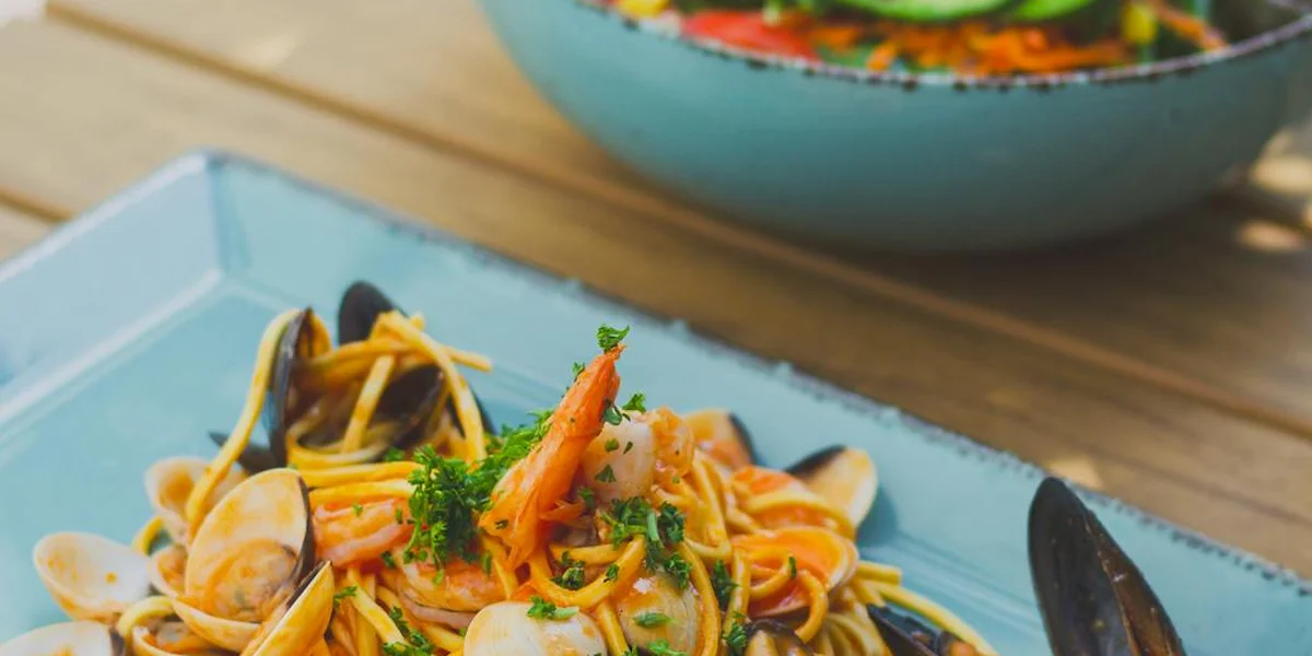 A blue plate of seafood pasta featuring shrimp, mussels, and herbs on a wooden table.