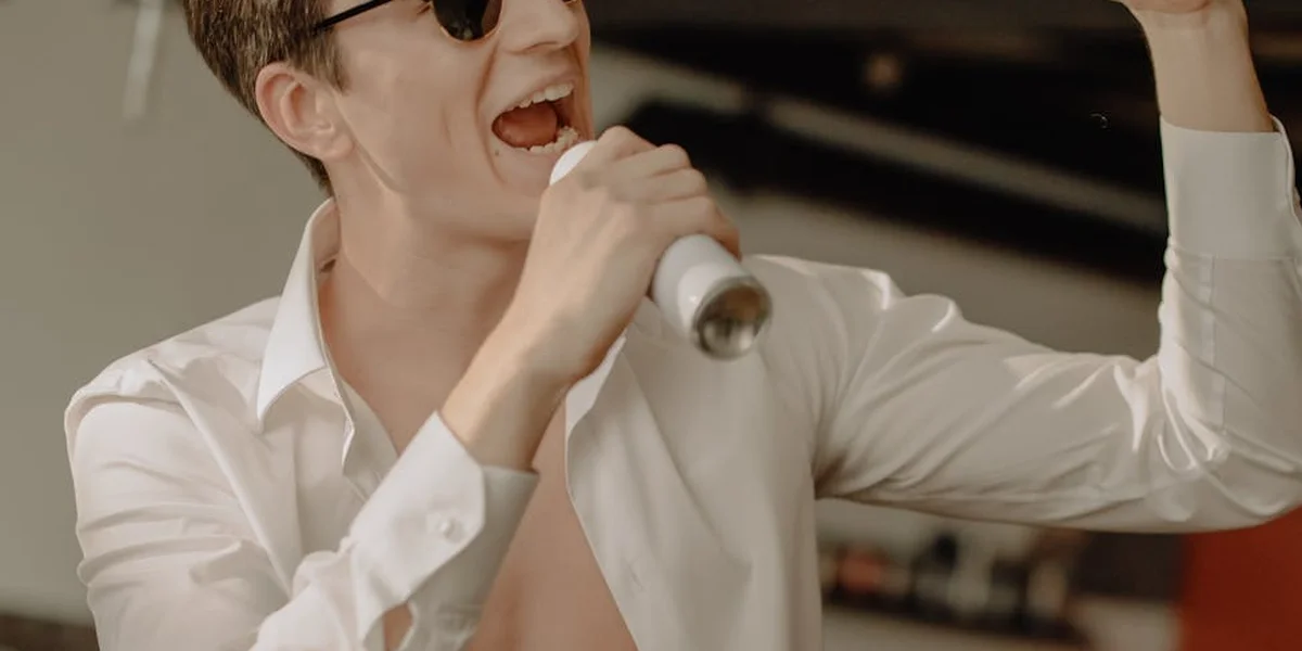 A person wearing a white shirt and sunglasses sings into a handheld microphone on stage, mid-performance.