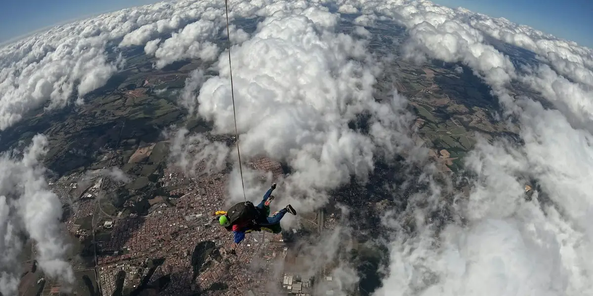 A skydiver is freefalling against a backdrop of scattered clouds and a patchwork landscape, with lines and equipment visible as they descend.