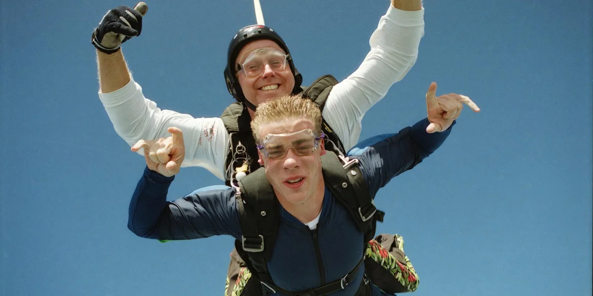 Two skydivers in tandem freefalling against a clear blue sky; the front diver smiles while the instructor behind him raises his arm.