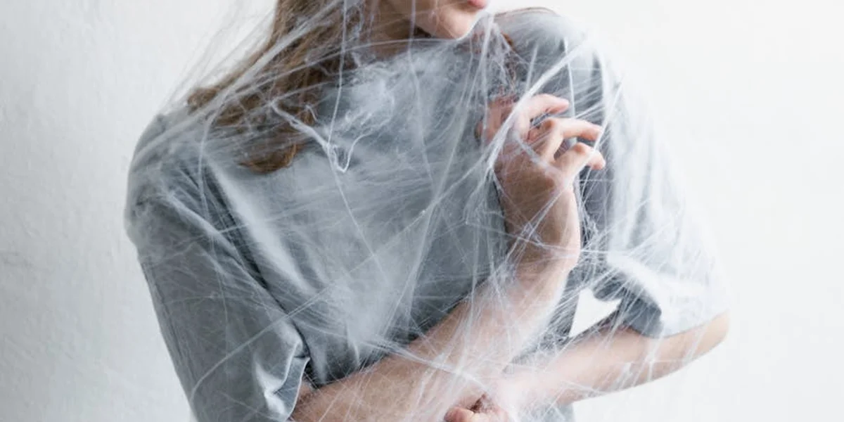 A person wearing a gray shirt is wrapped in translucent spider-web fabric, creating a dreamlike, entangled scene.