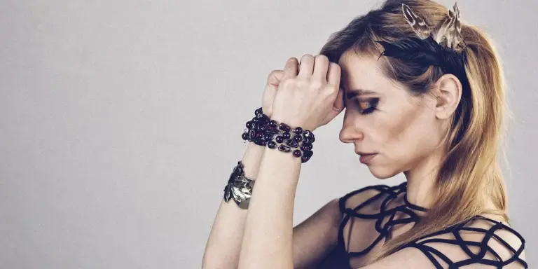 A pensive woman with a crown-like headband and multiple bracelets stands against a plain light background, her head slightly bowed.