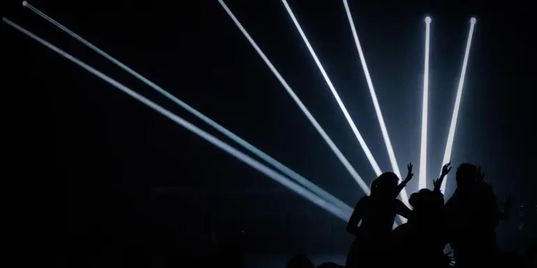 Silhouettes of performers on a dark stage, with dramatic white spotlight beams crisscrossing from above.