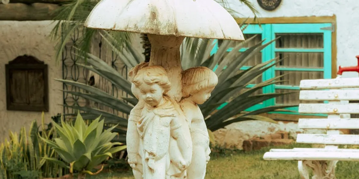 Two stone child statues sharing a large umbrella in a garden, with tropical plants and a turquoise window in the background.