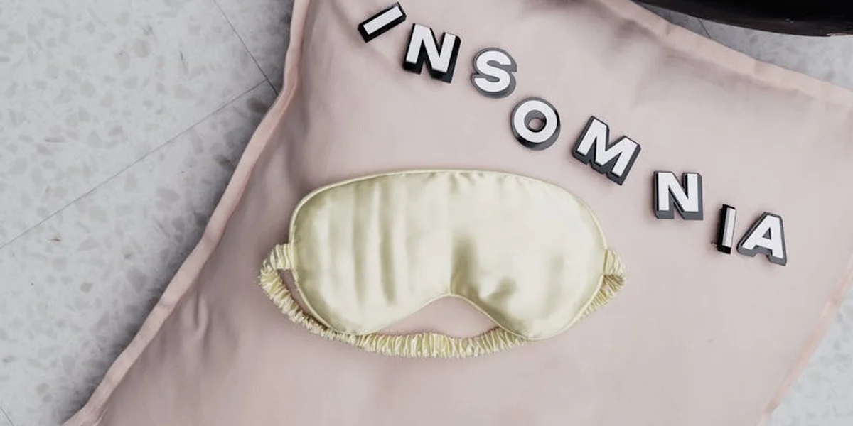 A pale sleep mask lies on a soft pink surface, with the word INSOMNIA spelled out in scattered letter tiles nearby.