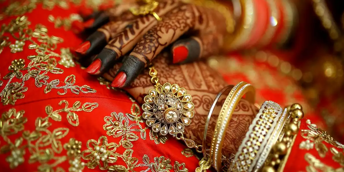 Close-up of hands with intricate henna patterns, adorned with gold jewelry and red fabric, suggesting ritual and spiritual symbolism.