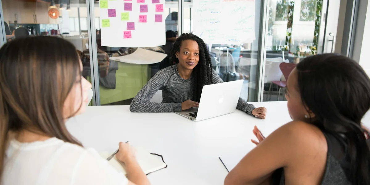 Three women seated around a bright conference table in a modern office; the woman in the center has a laptop and appears to lead the discussion while the others listen and take notes.