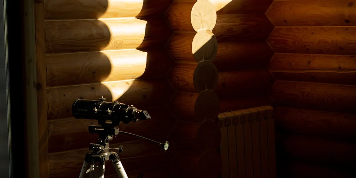 A telescope on a tripod inside a sunlit wooden log cabin, poised to explore from stars to tiny details.