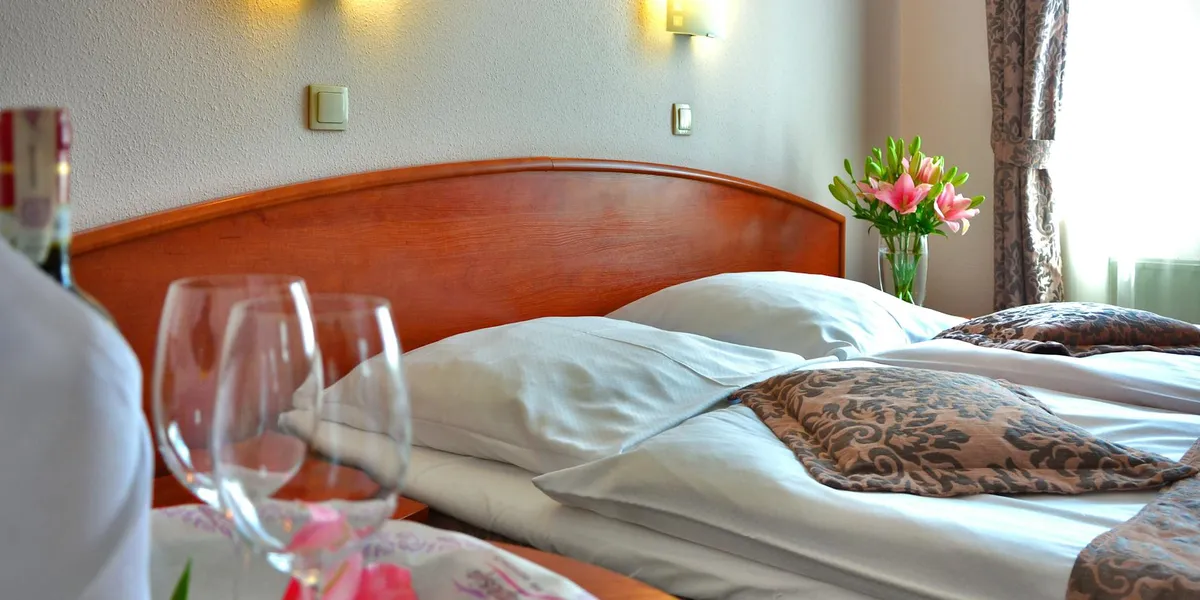 A tidy hotel bedroom with a wooden headboard, white bed linens, patterned pillows, a vase of pink flowers on the nightstand, and two wine glasses on a nearby table.
