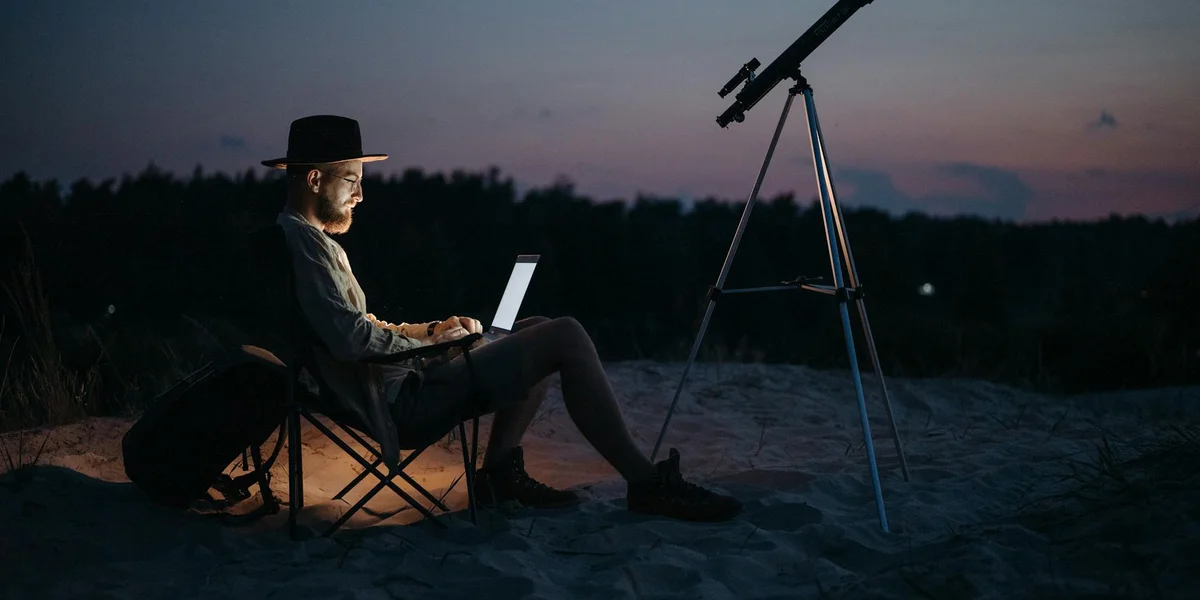 A person wearing a hat sits on a chair at dusk with a laptop on their lap, while a telescope on a tripod stands nearby.