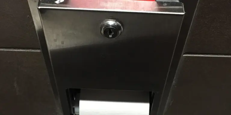 Close-up of a metal public bathroom fixture mounted on a dark tile wall, featuring a lock and a recessed slot.