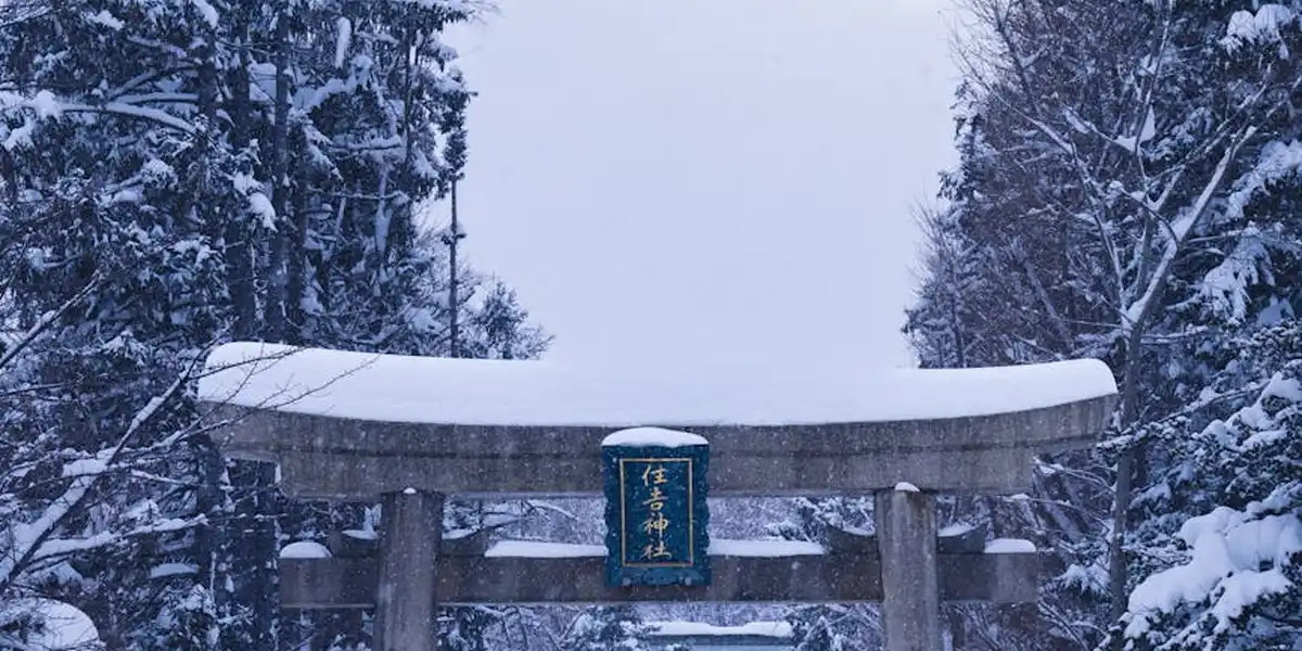 Snow-covered Shinto torii gate in a forest, blue plaque on the gate, with snow-laden pine trees in winter.