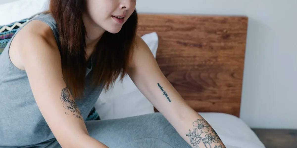 A person with long dark hair lying on a bed, wearing a sleeveless gray top, with tattoos visible on both arms.