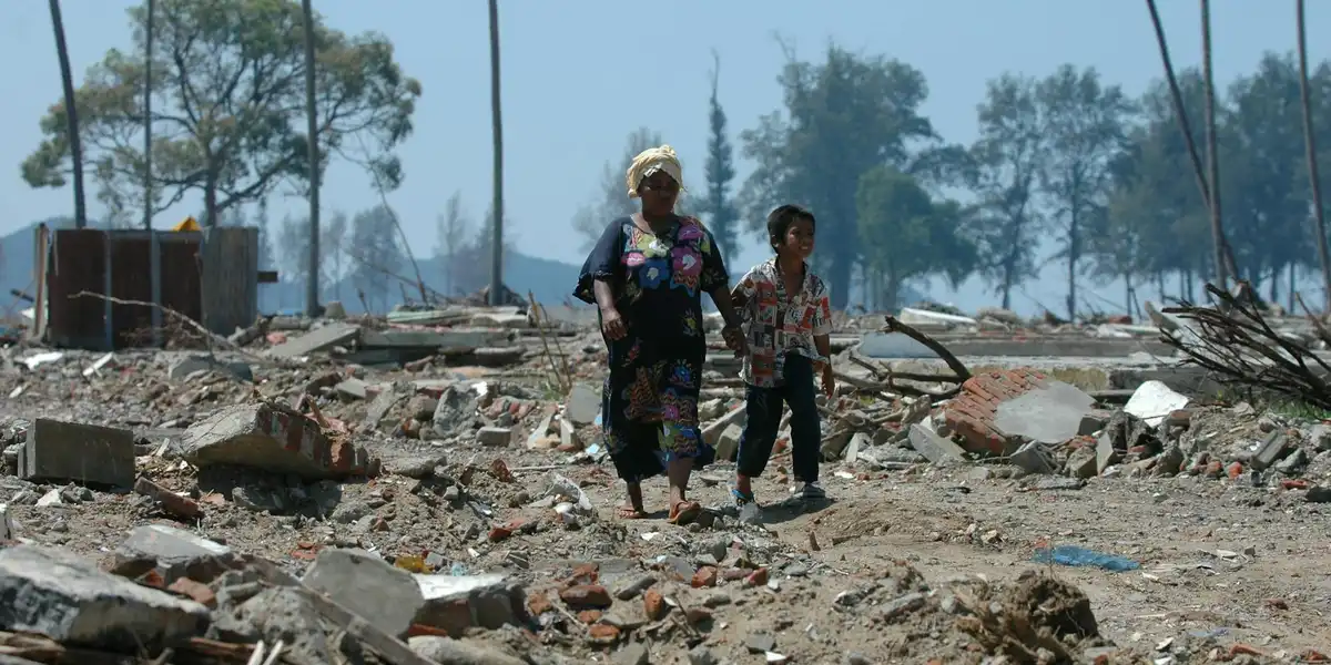Two people walk through a debris-strewn landscape with rubble, charred trees, and a hazy sky, depicting the aftermath of a disaster.
