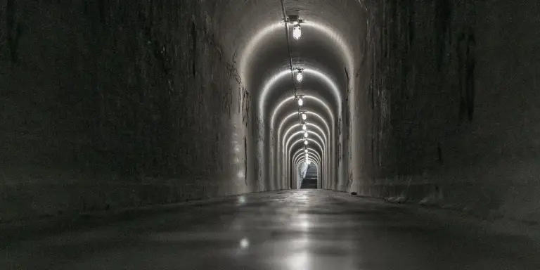 A long, dim tunnel with arched ceilings and a bright light at the far end, suggesting pursuit or escape.