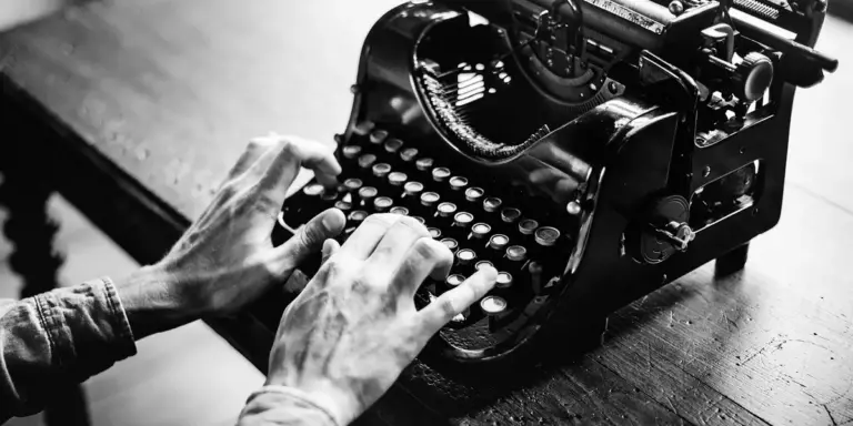 Black-and-white photograph of a pair of hands typing on a vintage manual typewriter on a wooden desk.