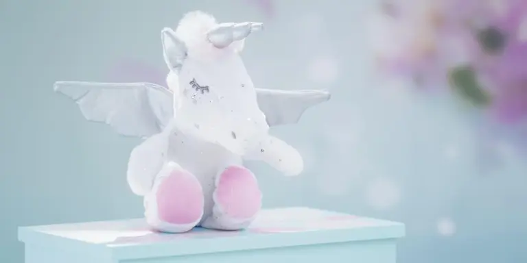 Soft white plush unicorn with pink hooves and tiny wings sits on a pastel blue background.