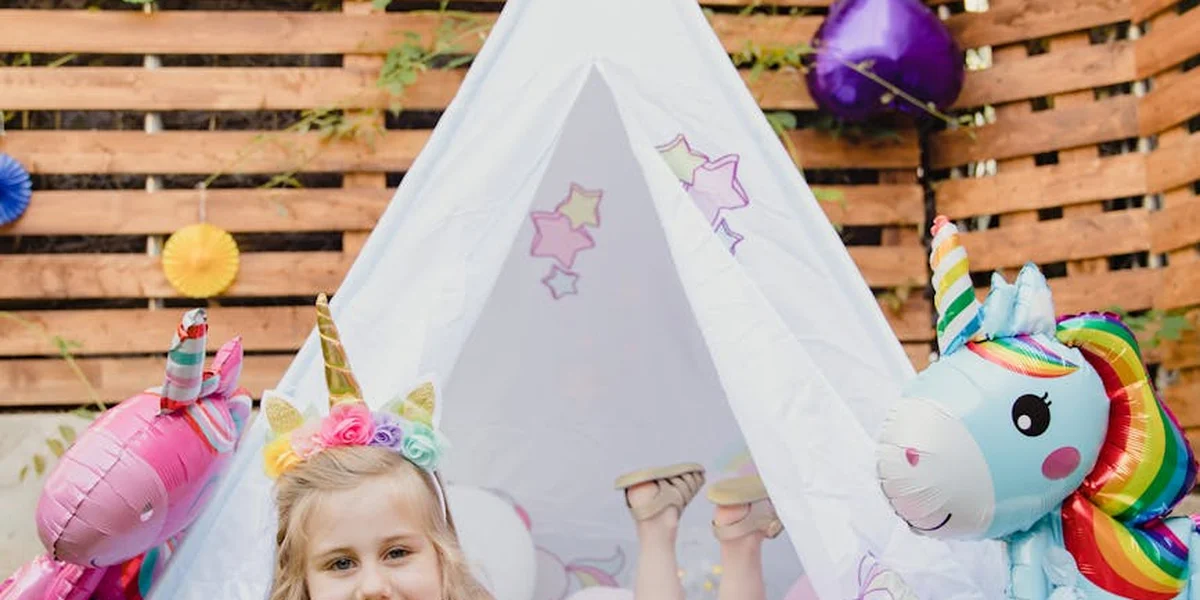 A child with a unicorn horn headband stands beside a white unicorn-themed tent and plush unicorn toys at a festive setup.