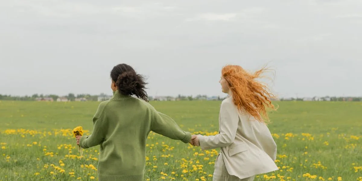 Two people hold hands while walking across a grassy field with yellow flowers, under a cloudy sky.