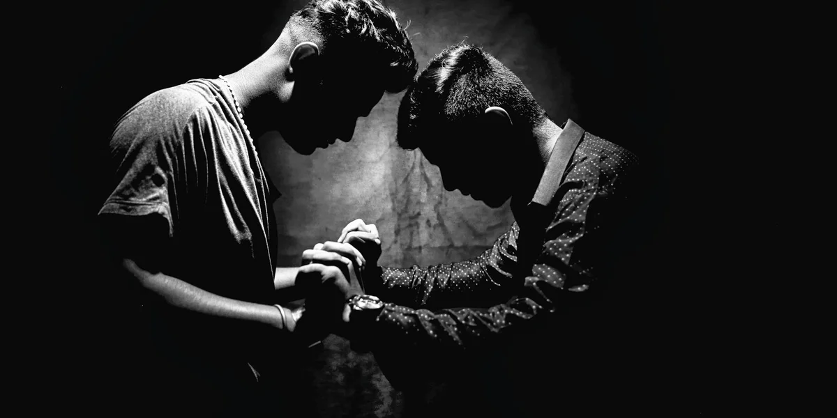 Two silhouetted figures lean toward each other, hands reaching to clasp in a dark, moody setting.