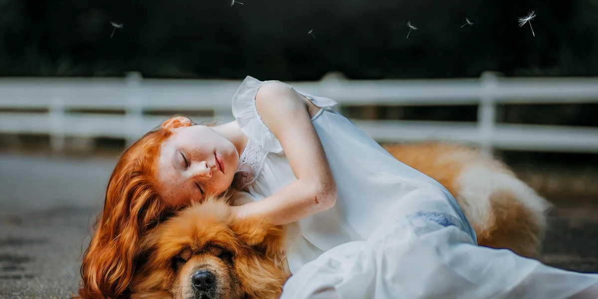 Person with long red hair lying on the ground beside a golden dog outdoors, near a white fence, suggesting comfort after loss.