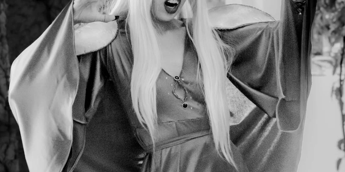Black-and-white portrait of a person wearing a vampire cape with long light hair, striking a dramatic pose evocative of vampiric imagery.