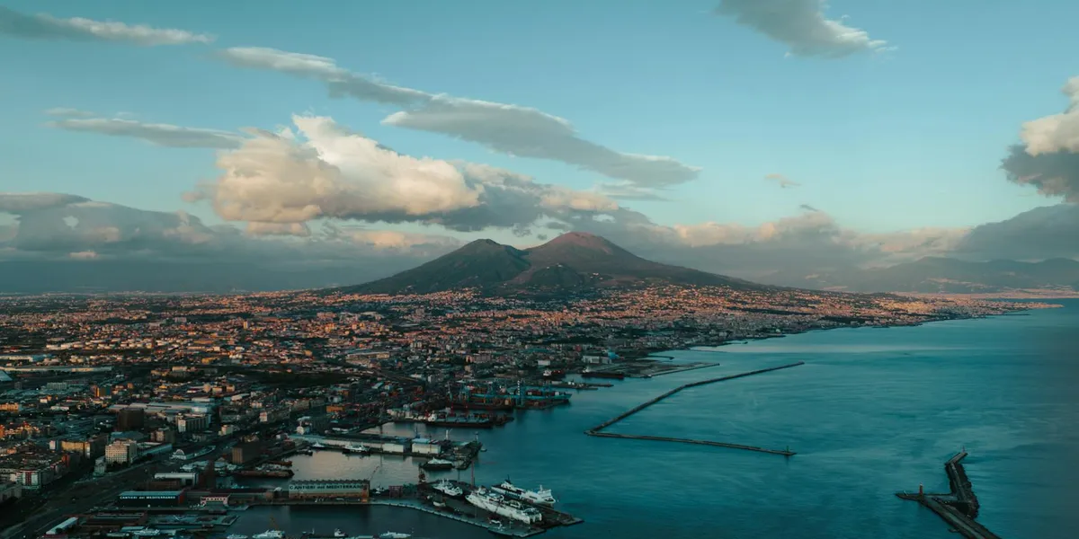 A distant, dormant volcano looms over a coastal city and turquoise sea under a bright sky.