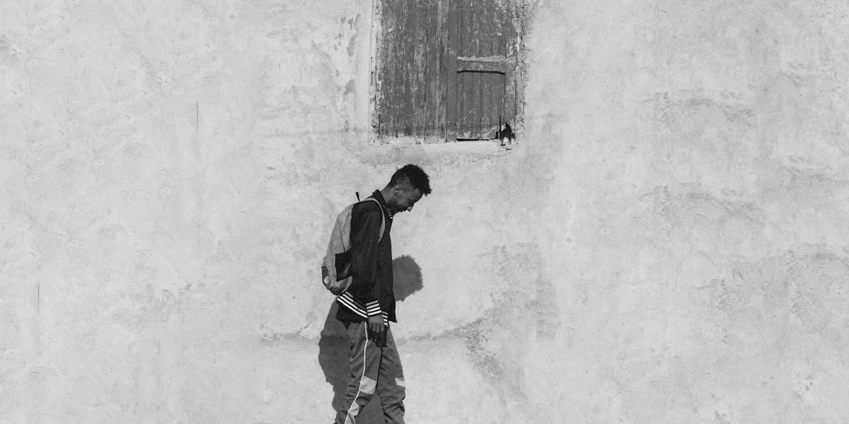 Black-and-white photo of a person walking along a textured wall with a small window above