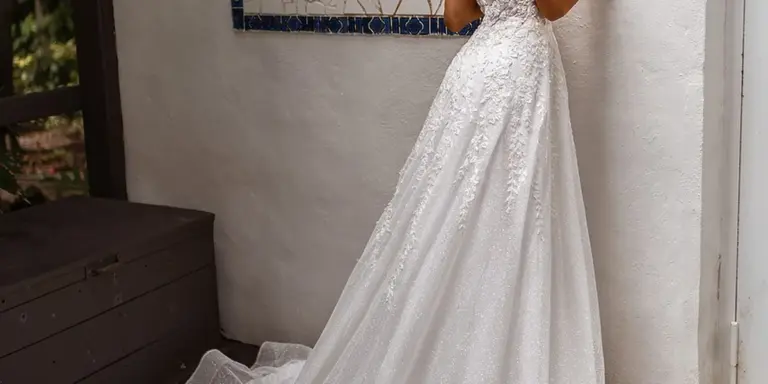 A white wedding dress with lace detailing worn by a person standing indoors, showing the full skirt and train against a neutral wall and window.