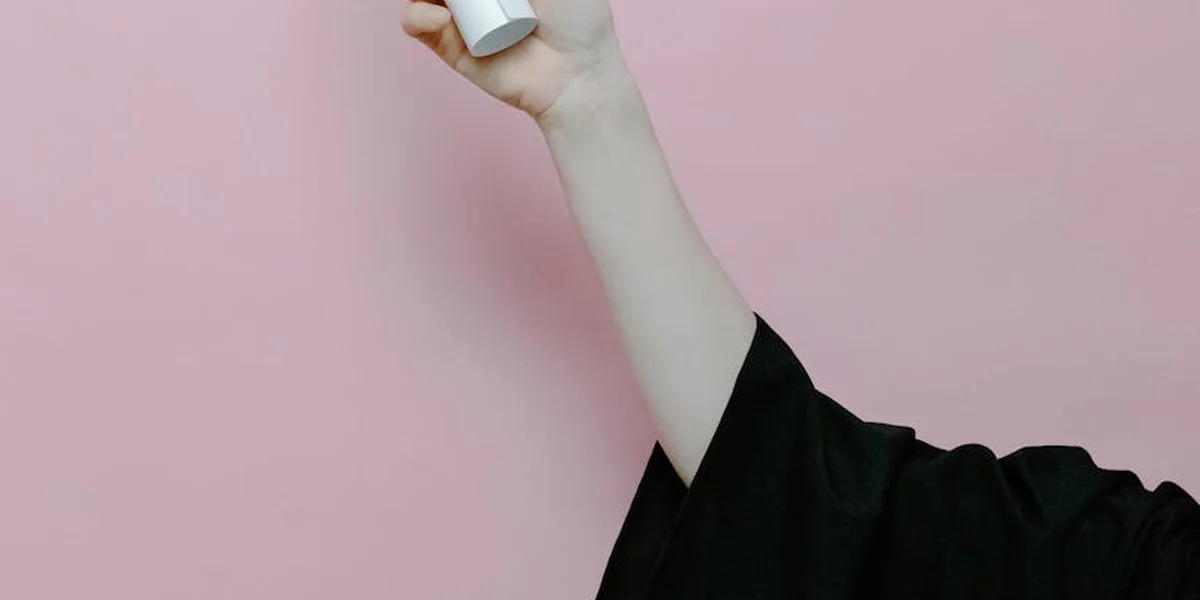 Close-up of a pale arm reaching upward, holding a small white object, set against a soft pink background.