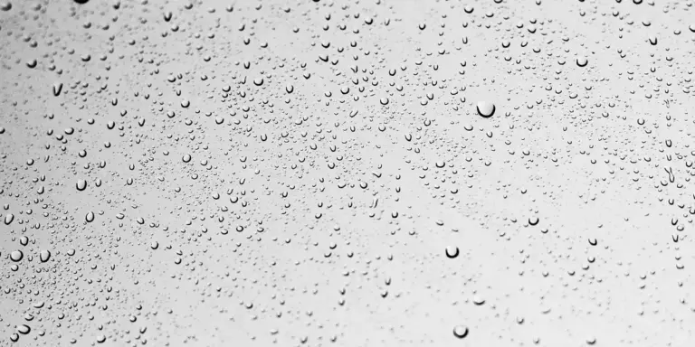 Close-up of raindrops on a glass window, with a soft blurred background in grayscale.