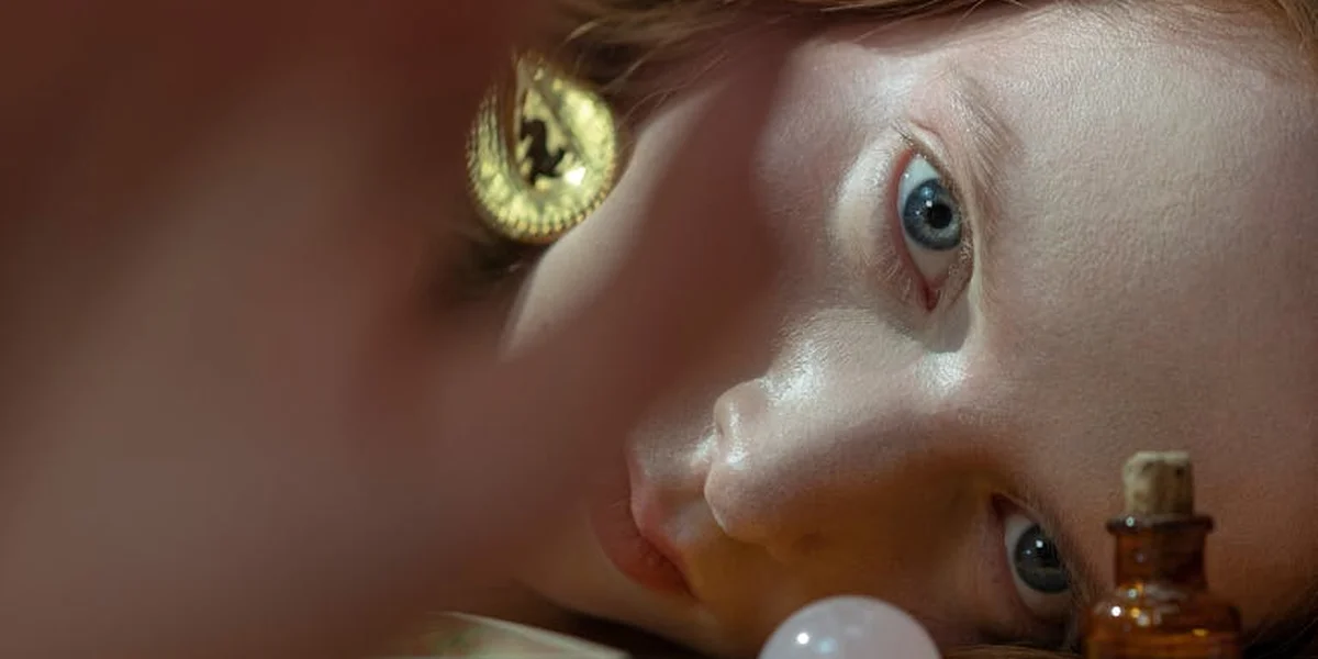 Close-up of a pale-skinned person with light blue eyes, holding a small potion bottle, with a gold pendant visible in the foreground.