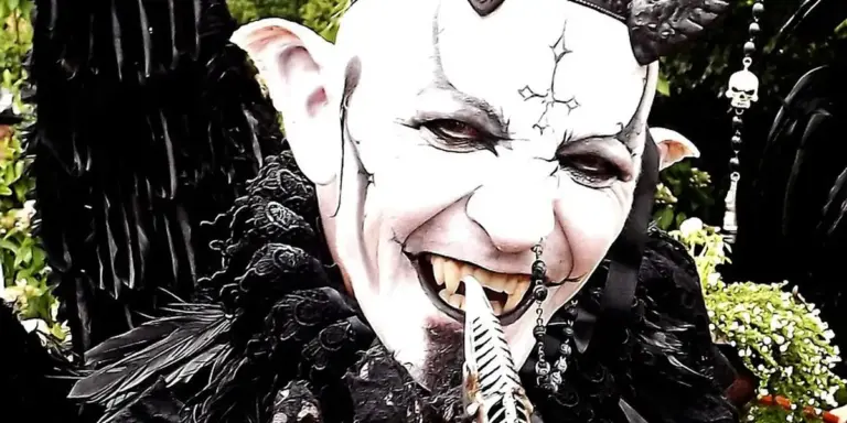 A costumed figure in a black cloak with pale makeup, horn-like headpiece, and eerie smile.