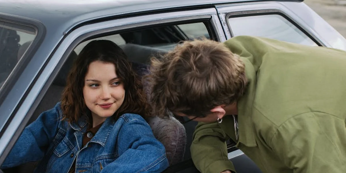 Two young people sit in a car; the woman in a denim jacket smiles softly while the other person in a green hoodie leans toward her, creating a moment of emotional tension.