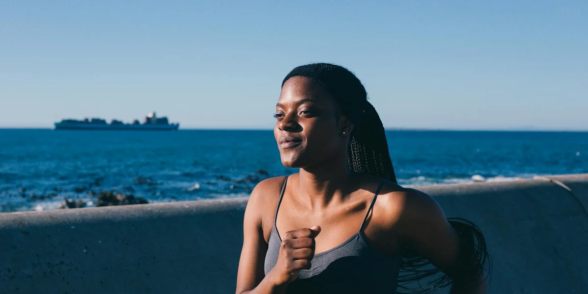 A woman stands by a sea wall, gazing off to the side with a calm expression, wearing a gray tank top, with the ocean and a distant ship in the background.