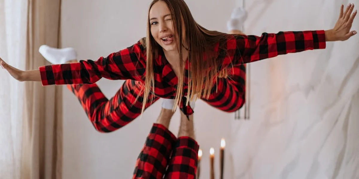 Woman in red and black plaid pajamas performing a balancing pose indoors, with candles in the background.