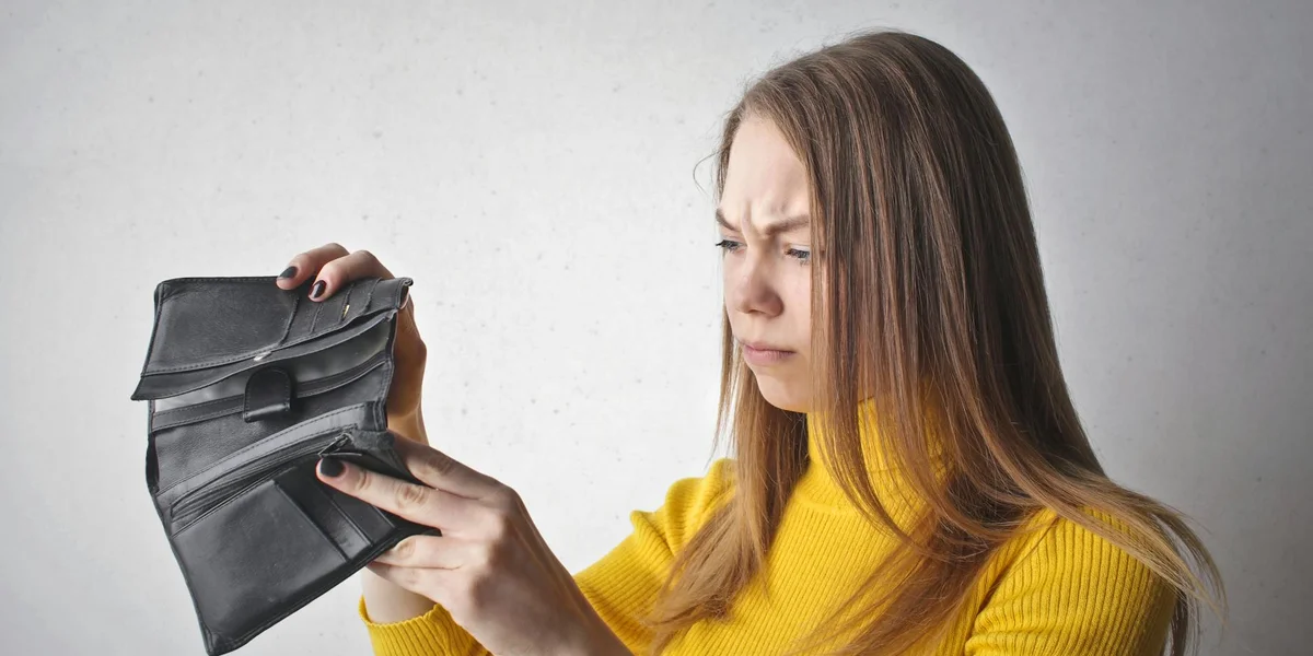 A woman wearing a yellow sweater stands against a light gray backdrop, examining an open black wallet with a concerned expression.