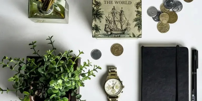 Top-down view of a tidy desk with a potted plant, a wristwatch, coins, a black notebook, a pen, and a vintage world map box.