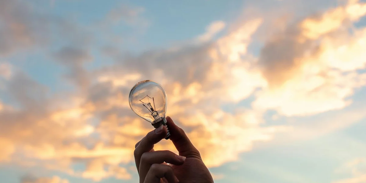 Close-up of a hand holding a glowing lightbulb against a warm sunset sky with clouds.