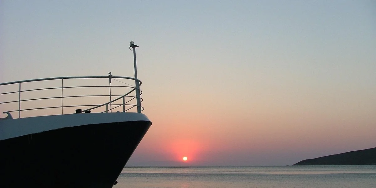 Sunset over the sea with the bow of a ship on the left and a warm yellowish-orange sky.